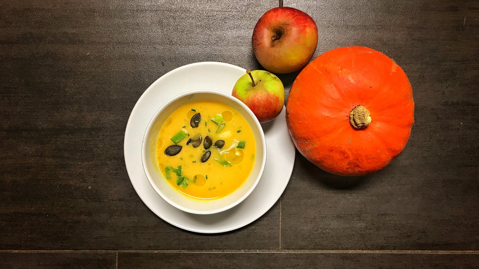 Cremesuppe vom Kürbis mit gerösteten Kürbiskernen, daneben ein Hokkaido-Kürbis und Äpfel